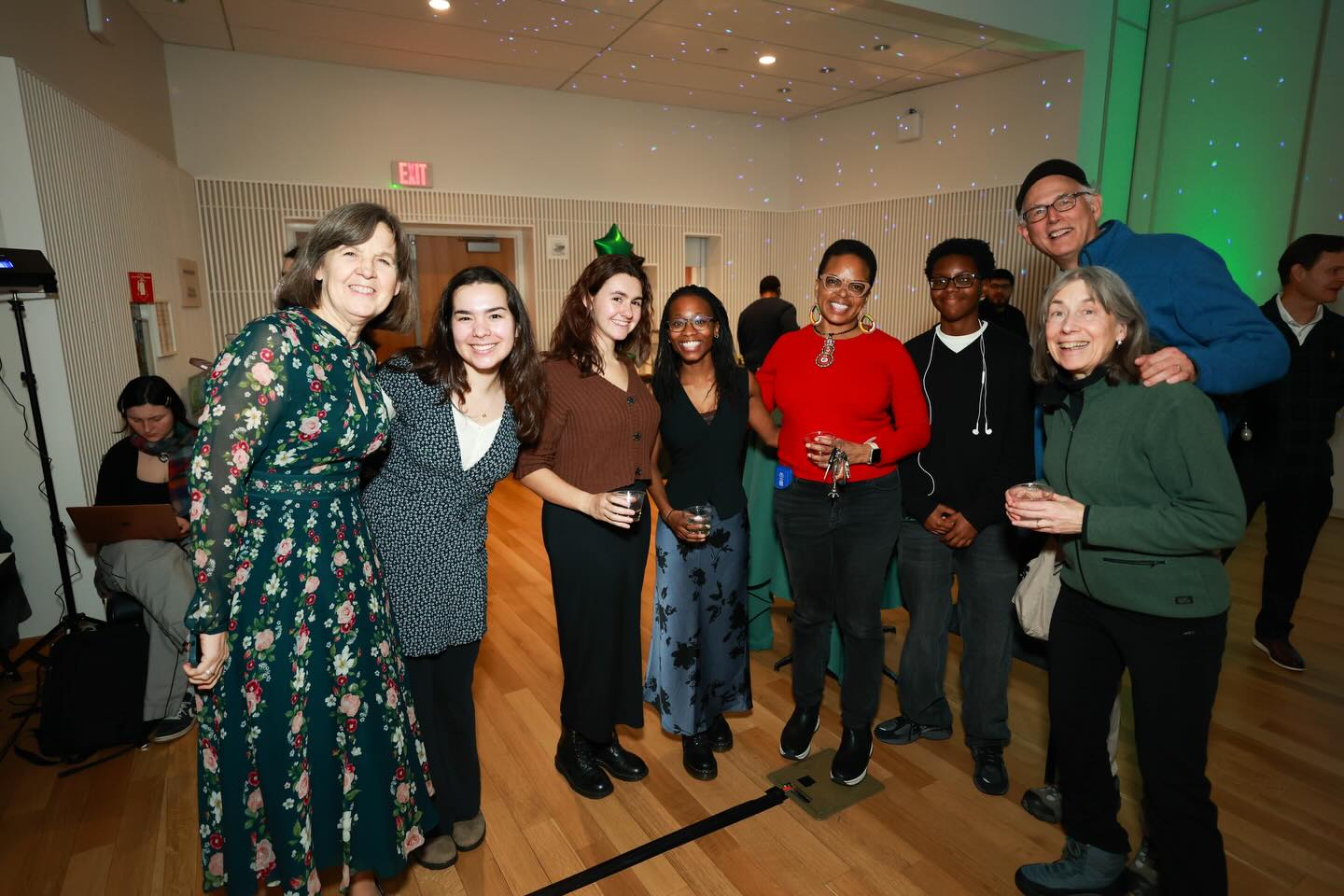 A group of people smiling at a casual indoor event, with some holding drinks and wearing a mix of colorful and neutral outfits.