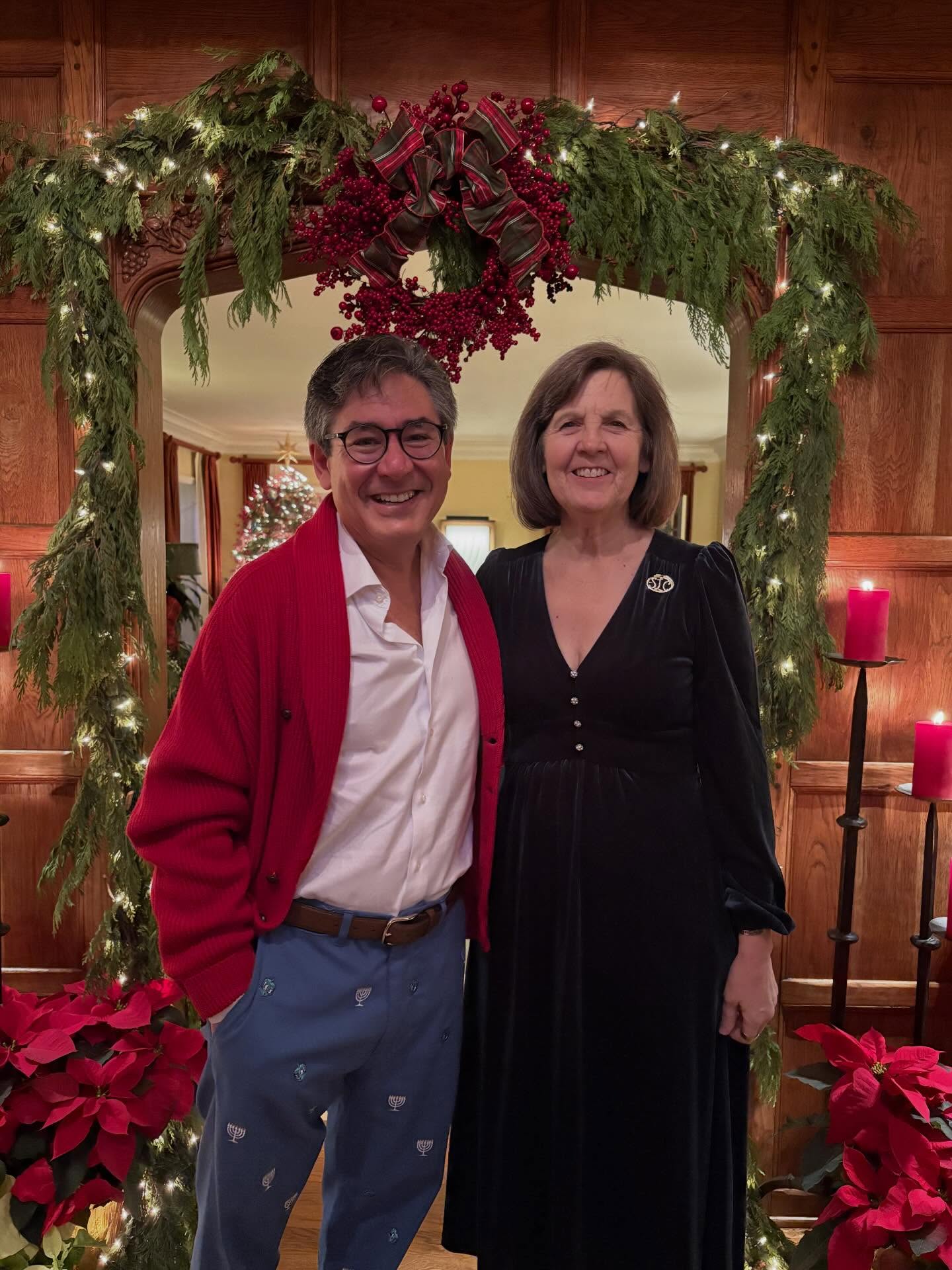 Two people smiling in front of a festive wreath and garland with poinsettias and candles, one wearing a red cardigan, the other in black attire.