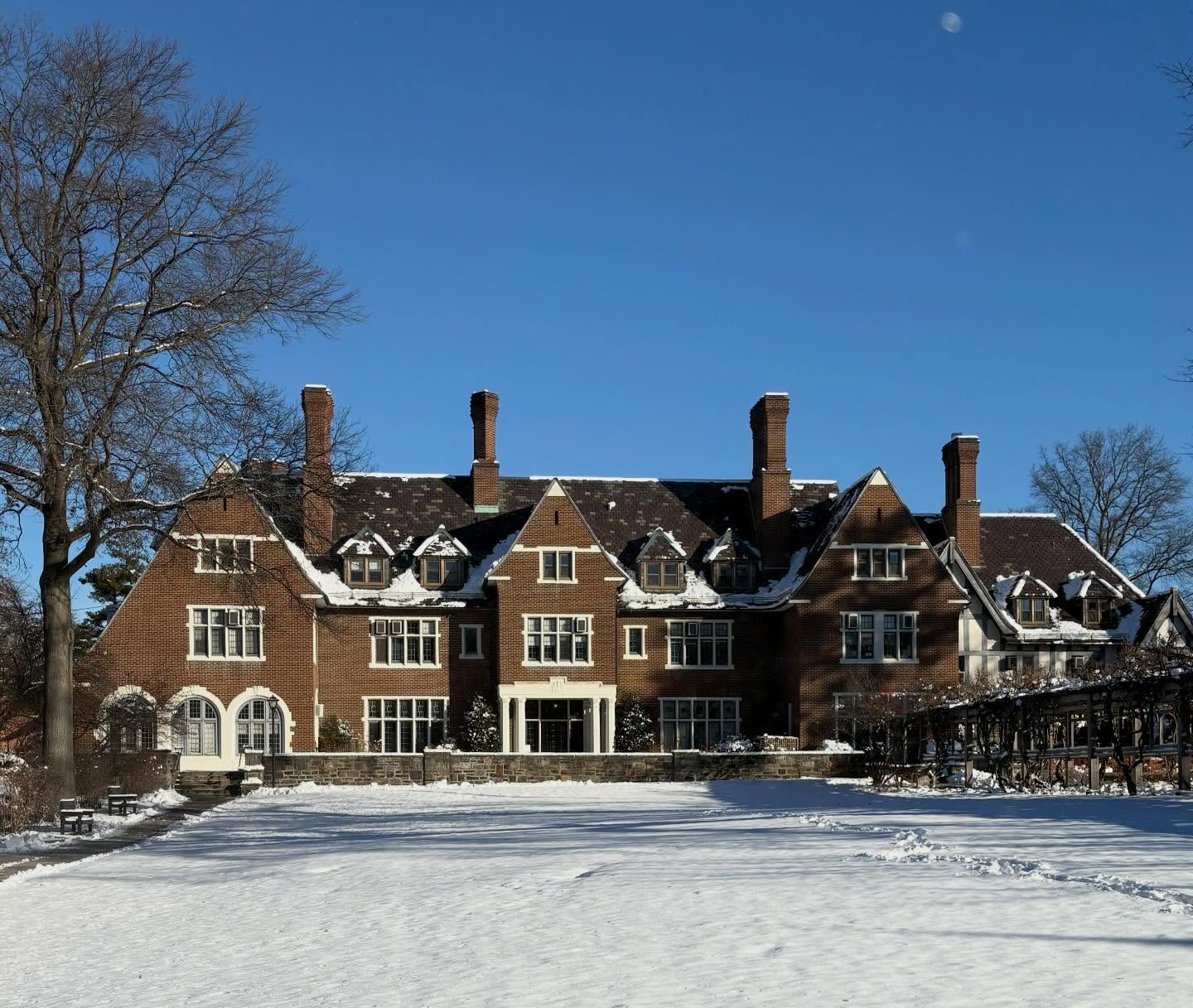 Large brick mansion with multiple chimneys and snow-covered roof, set against a clear blue sky. Snowy lawn in the foreground.