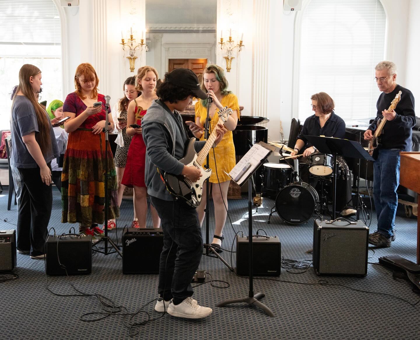 Group of musicians performing indoors, featuring guitarists, vocalists, and a drummer. A standing audience observed nearby.