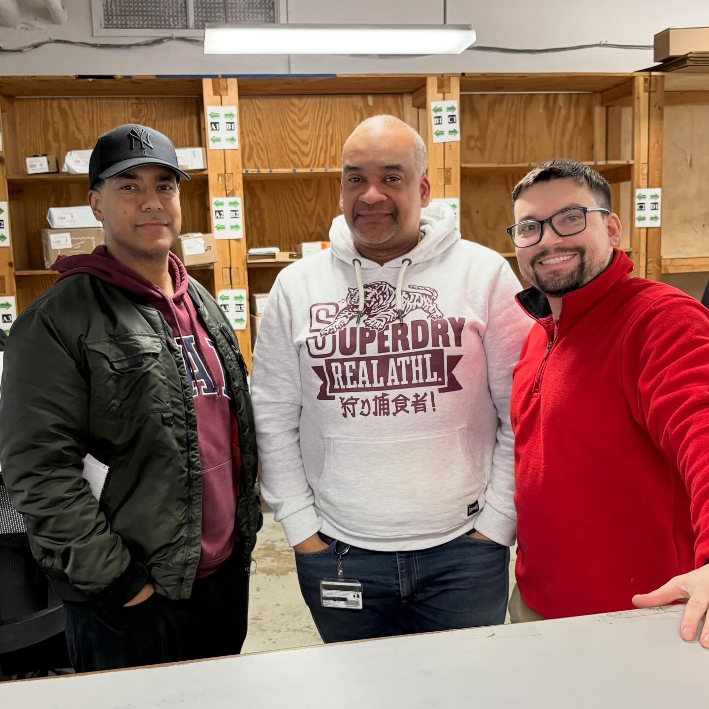Three people stand in a room with wooden shelves behind them, wearing casual hoodies and jackets, smiling at the camera.