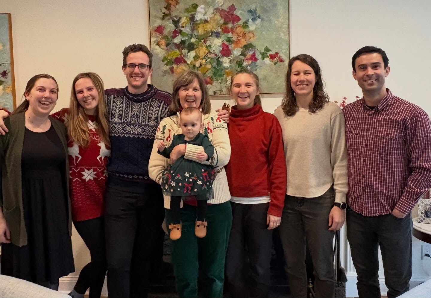 Seven adults and one child in festive sweaters stand smiling indoors, with a colorful painting in the background.