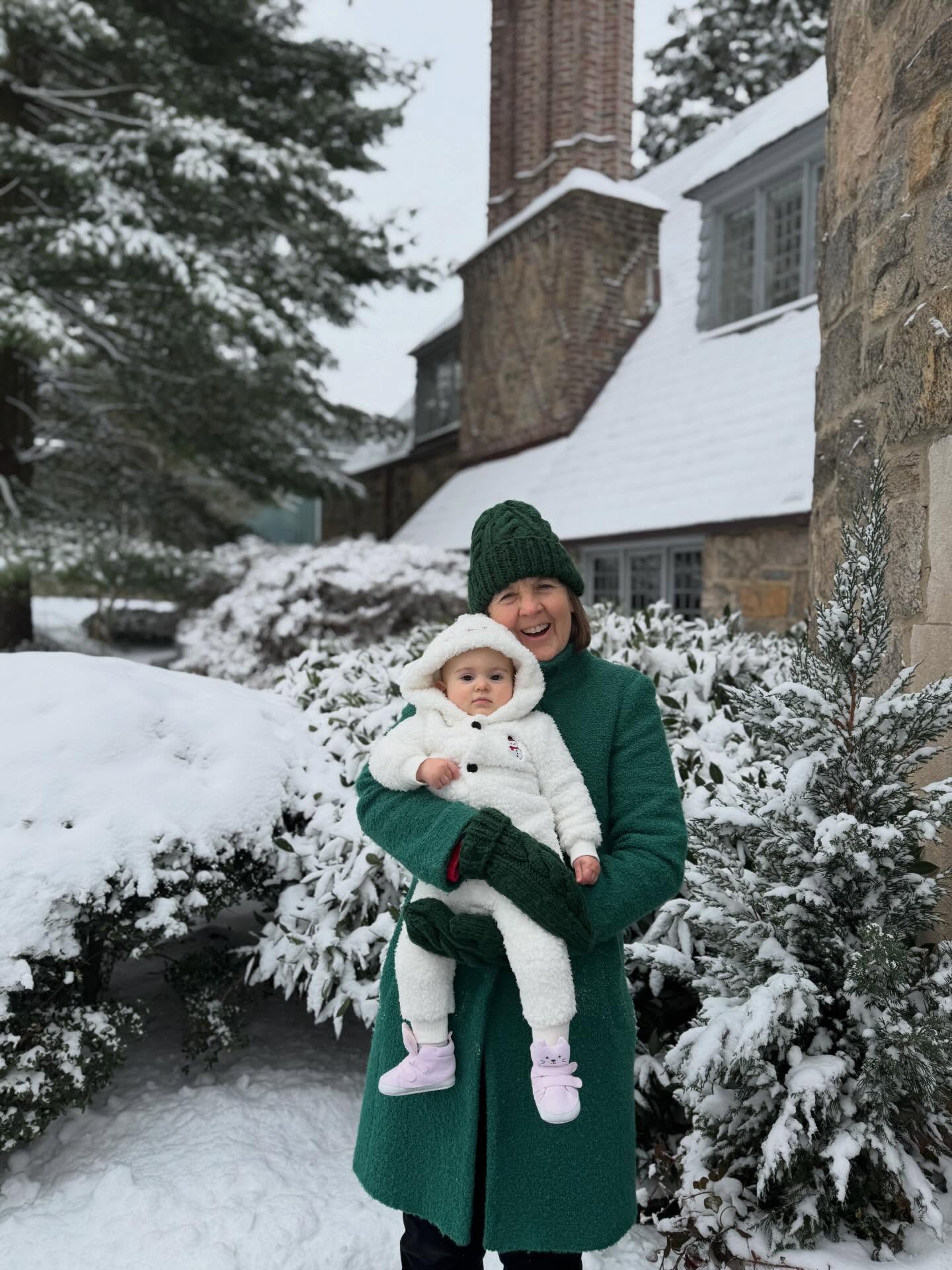 Person in a green coat and hat holds a baby in a fluffy white snowsuit. Snowy background with a large stone house and trees.