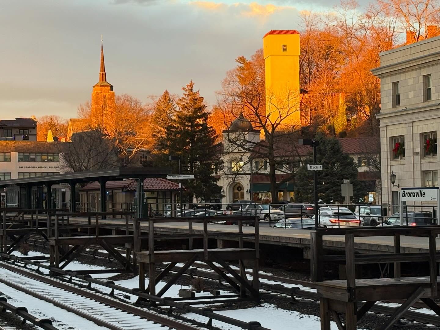 Train tracks with snow in foreground, a platform with cars and buildings, including a tall church tower and orange-lit structure in the background.