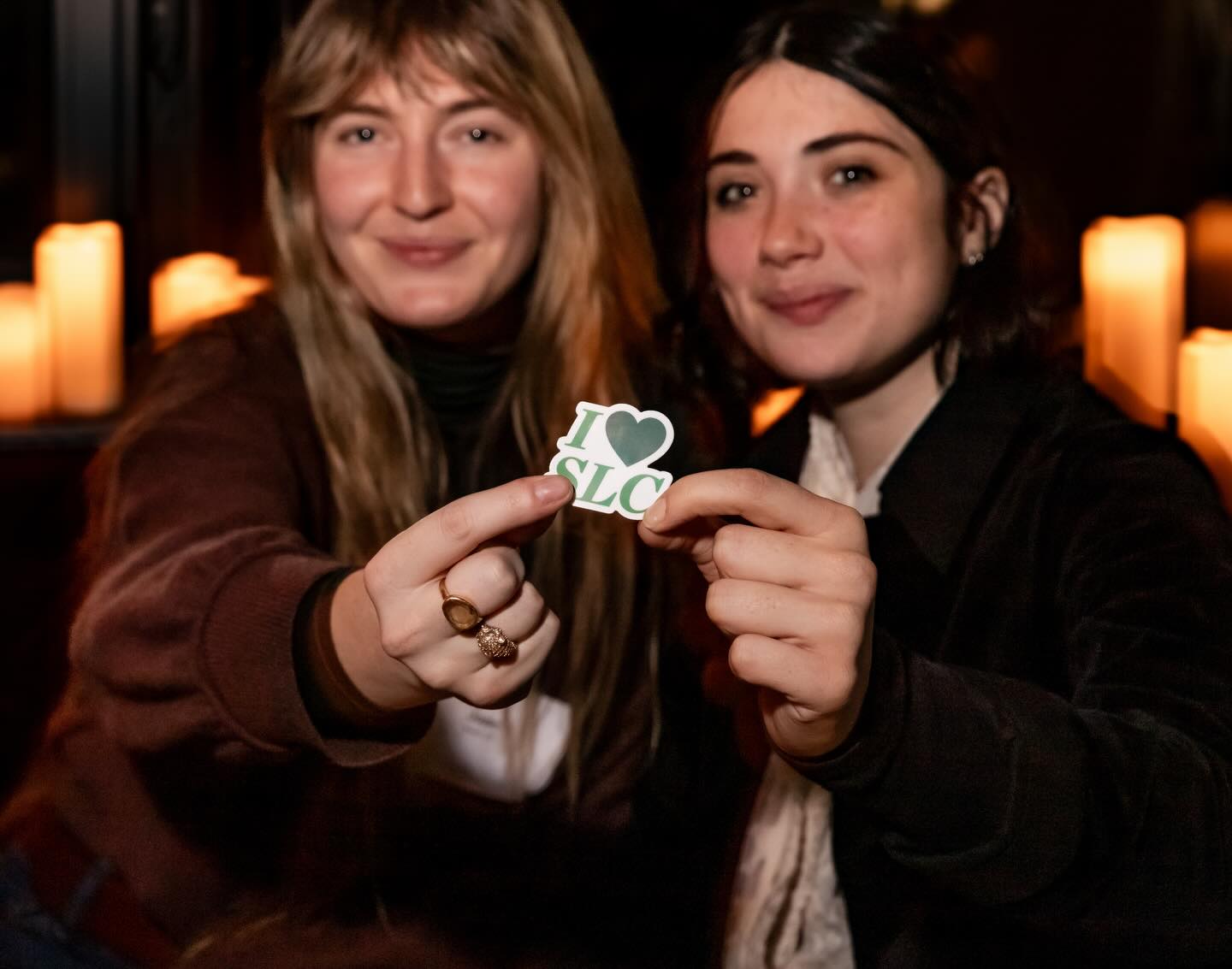 Two people holding a sticker that says "I ♥ SLC" with candles glowing in the background.
