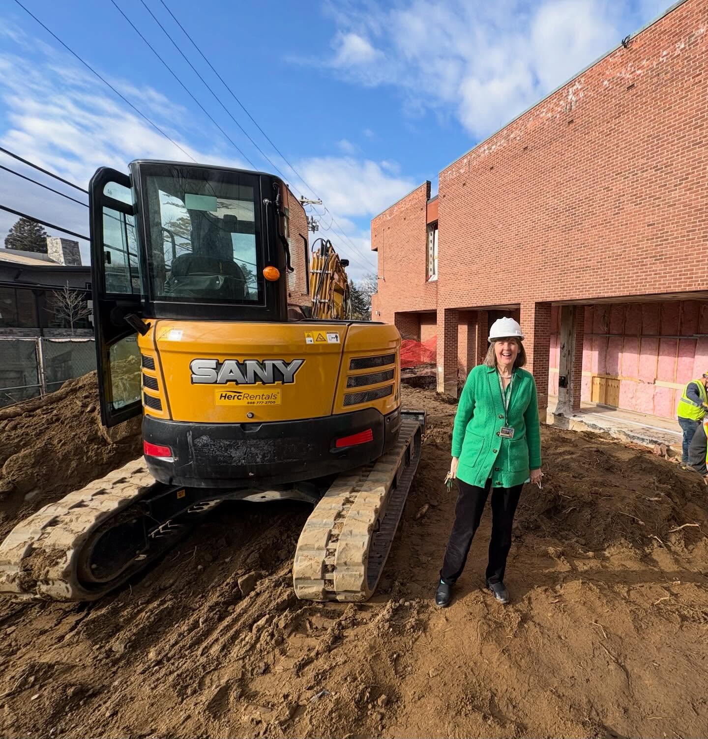 Person wearing a green jacket and white helmet stands next to a yellow excavator on a construction site with red-brick building in the background.