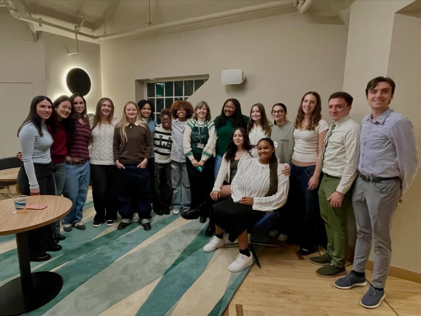 A group of 16 people smiling indoors, standing and sitting in a well-lit room with a light-colored wall and patterned carpet.