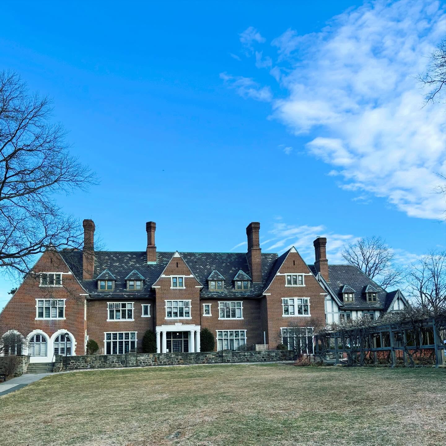 Large brick mansion with multiple chimneys and gables against a clear blue sky, surrounded by leafless trees and a grassy lawn.
