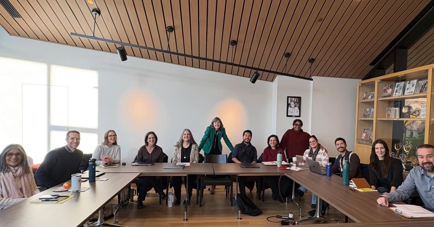Group of people sitting and standing around a large conference table, smiling, with papers, notebooks, and laptops present. Wooden ceiling and shelves in background.
