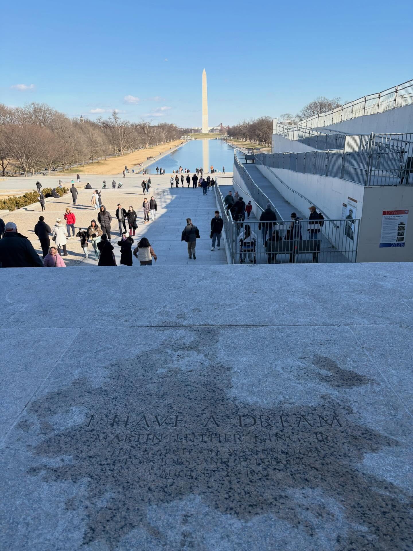Steps with engraved text lead to a reflecting pool, with the Washington Monument and people walking in the distance.