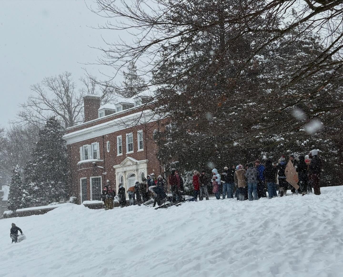 A group of people in winter clothing stand in front of a brick building during snowfall, surrounded by snow-covered trees.