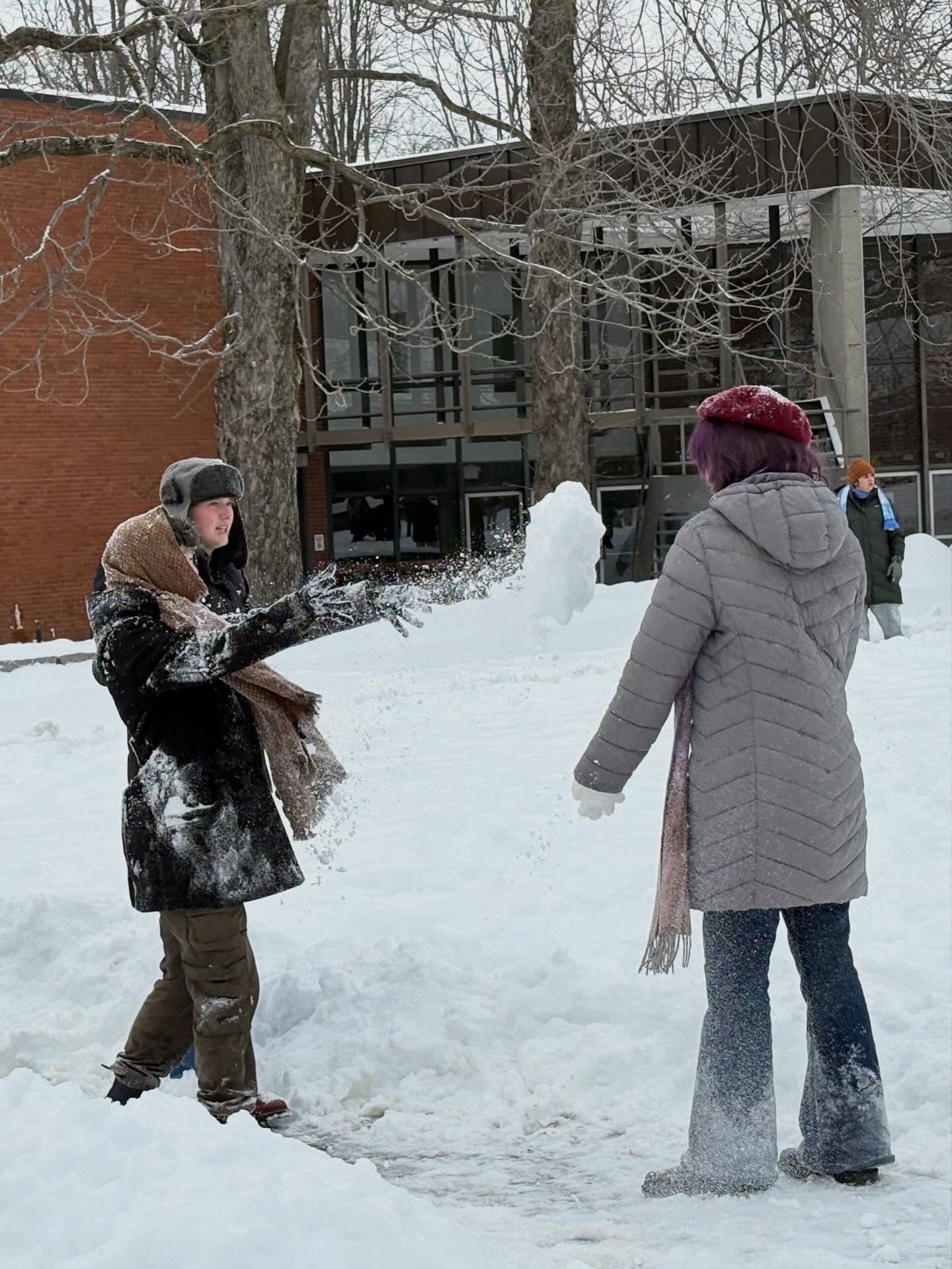 Two people having a snowball fight near a building, with snow-covered ground and trees in the background. Another person walks away.