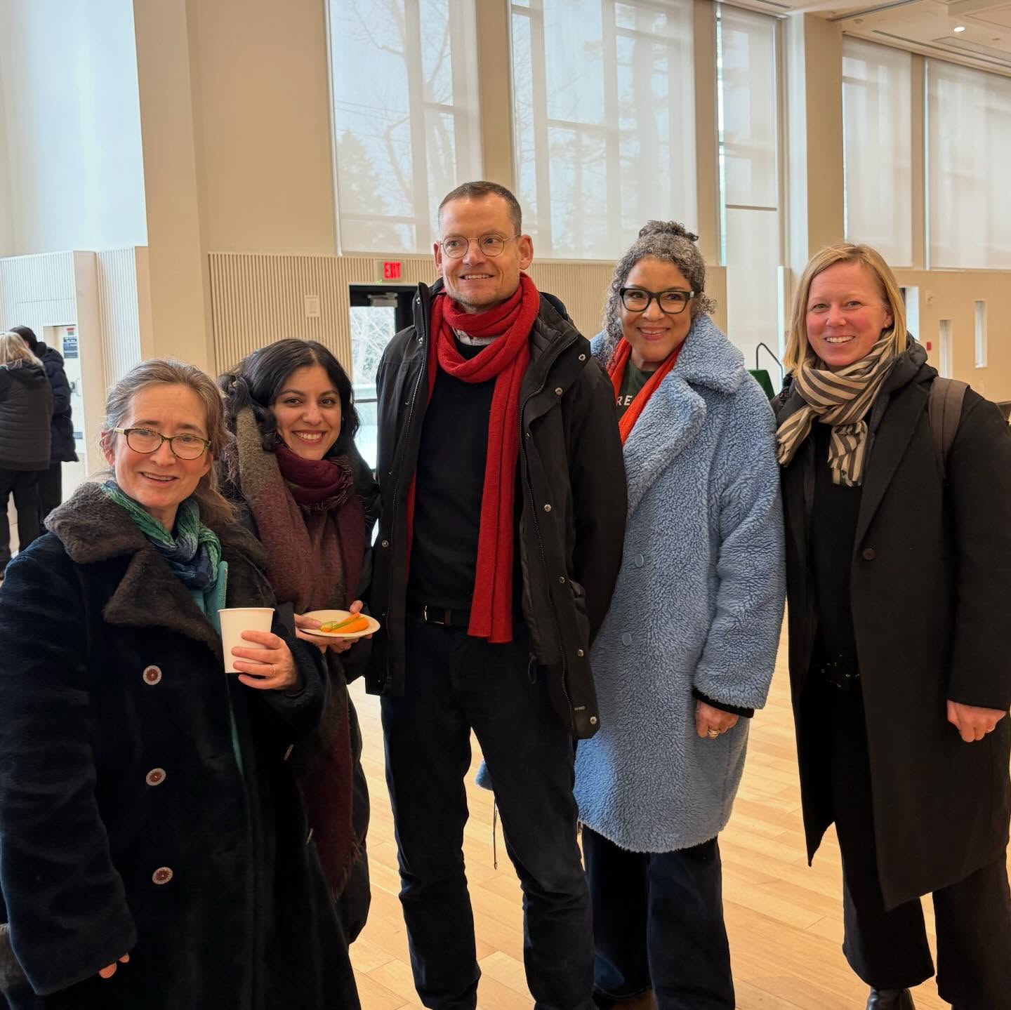 Five people in winter clothing smiling indoors with large windows in the background. One holds a cup, another holds a plate with food.