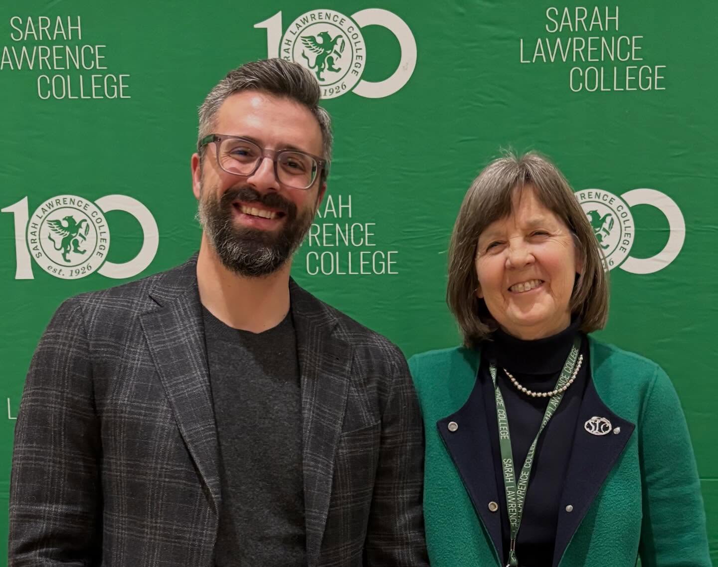 Two people smiling in front of a green backdrop with "Sarah Lawrence College" text and logo, celebrating its 100th anniversary.