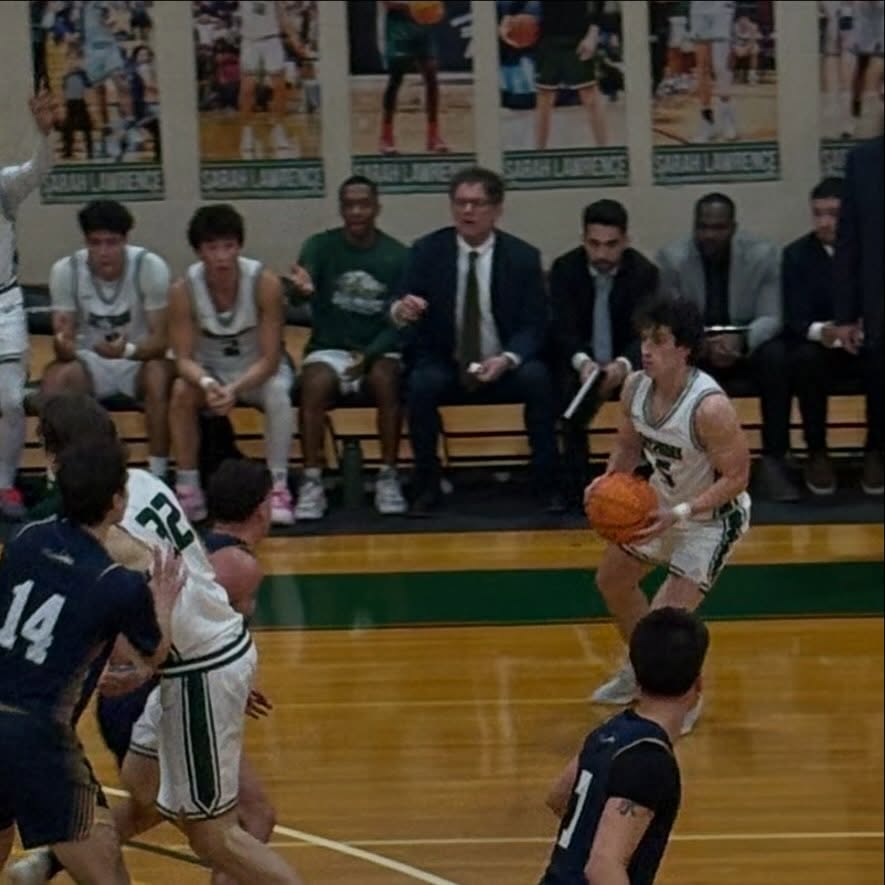 Basketball game in progress, player in white dribbling on court, others in action. Coaches and team in the background on the bench.