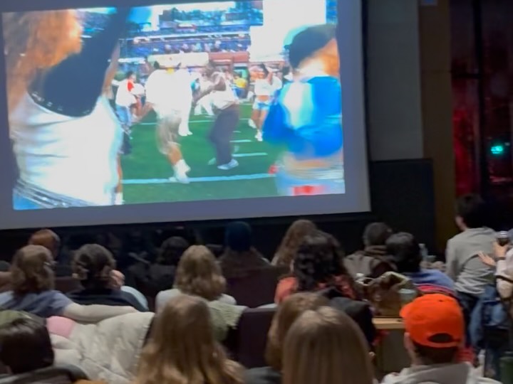 Audience seated in a room watching a sports event on a large screen showing a field and performers.
