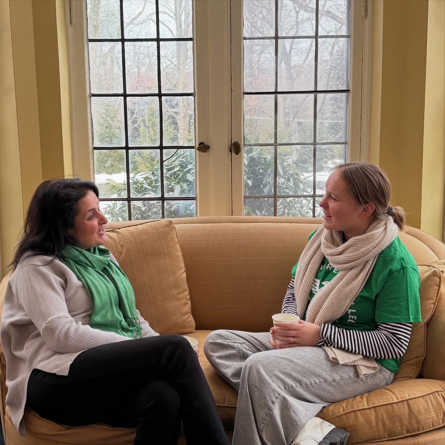 Two people sitting on a tan couch in a cozy room with large windows, chatting with cups in hand.