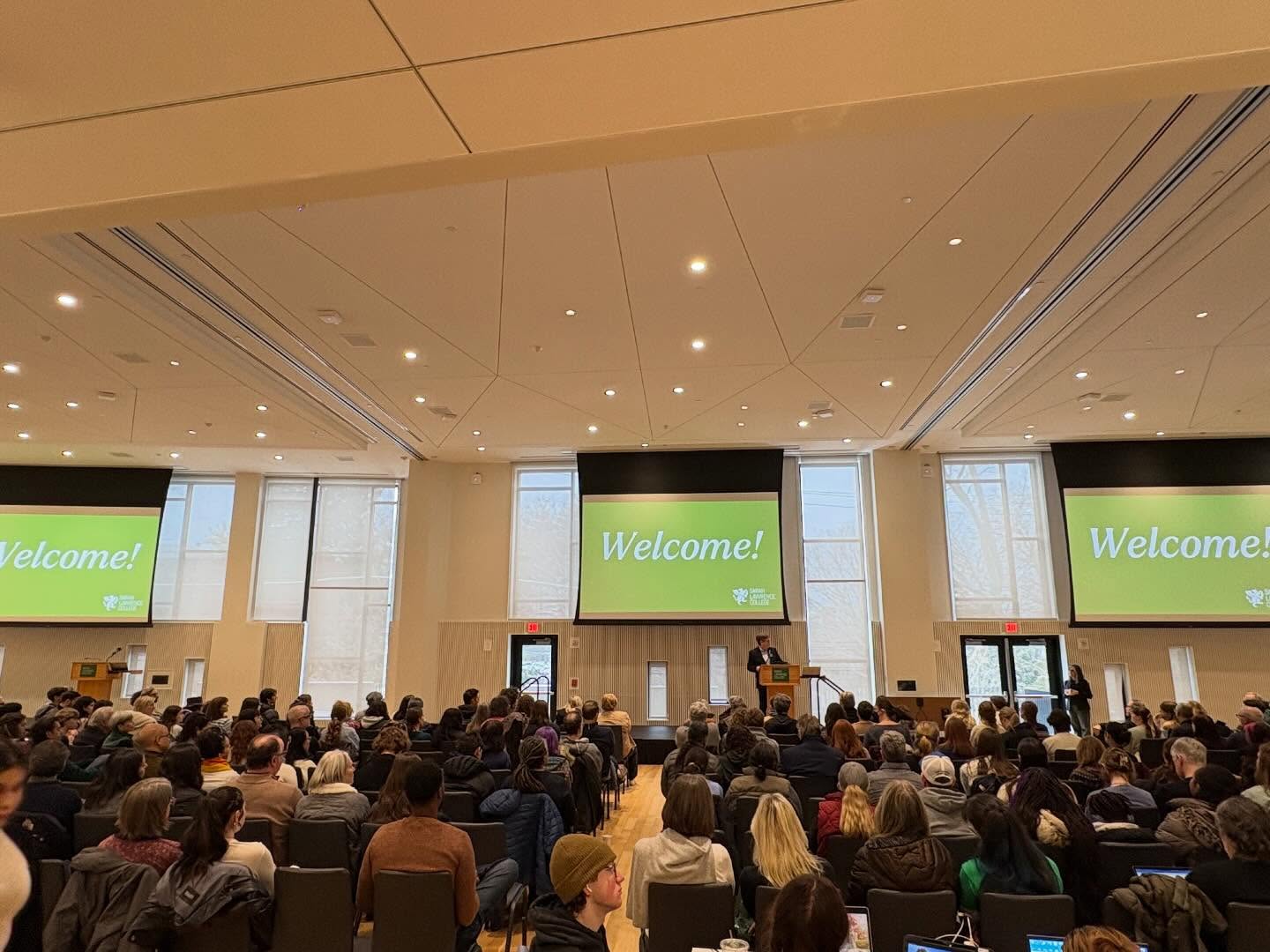 Audience seated in a large room facing a stage with a "Welcome!" message displayed on multiple screens.