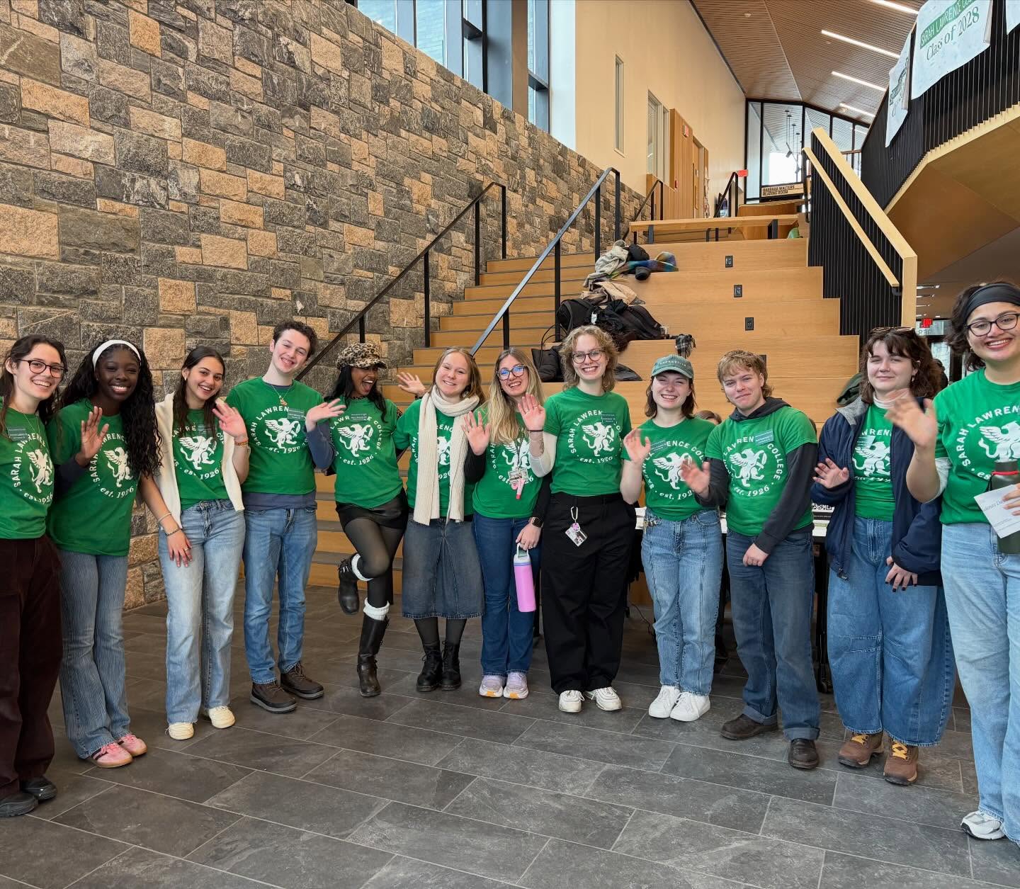 Group of people in matching green T-shirts smiling and waving in a building with a stone wall and staircase in the background.
