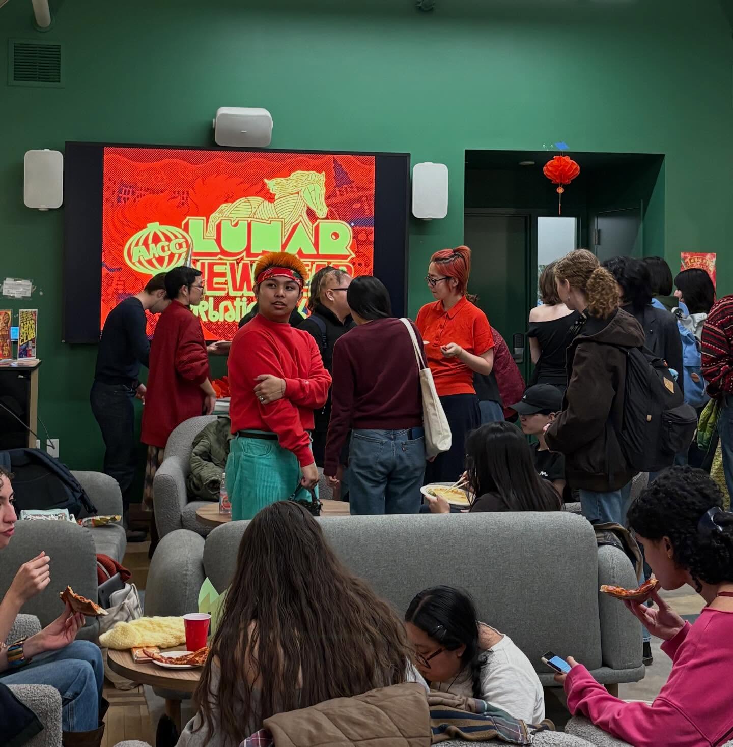 Crowd celebrating Lunar New Year indoors. People are talking, eating pizza, and gathered around a screen with festive decorations.