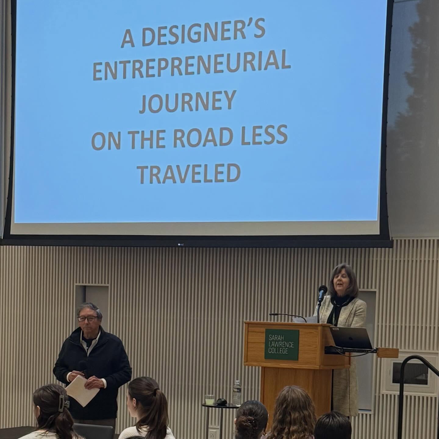 Two presenters stand in front of an audience. The screen shows "A Designer's Entrepreneurial Journey on the Road Less Traveled."