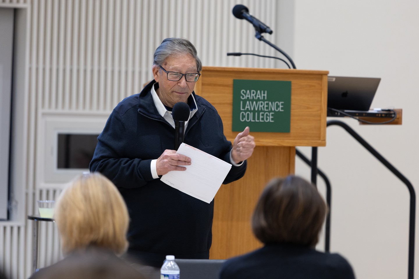 Person speaking at a podium with a microphone, holding papers at Sarah Lawrence College, facing an audience.