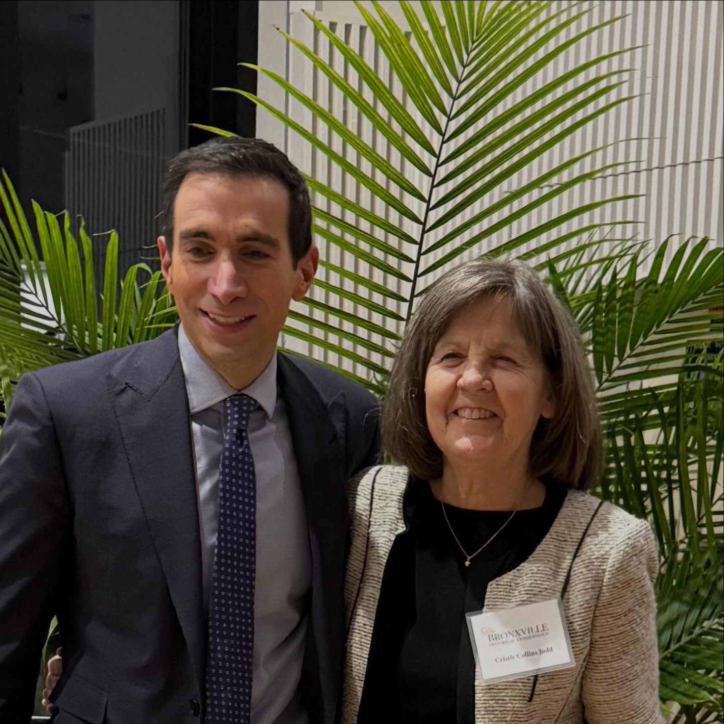 Two people smiling, one wearing a suit and tie, the other with shoulder-length hair and a nametag, standing in front of large green plants.
