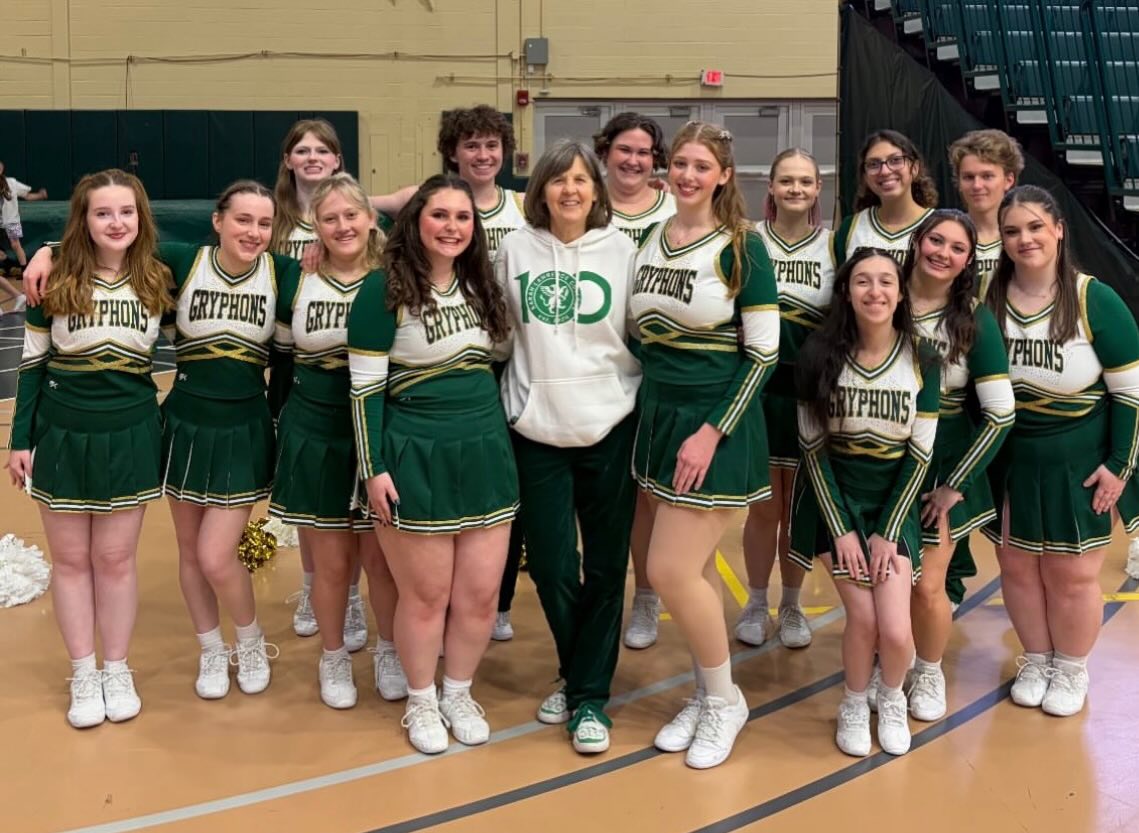 Cheer squad in green and white uniforms with "GRYPHONS" across the chest, posing with a person in a white hoodie in a gym.