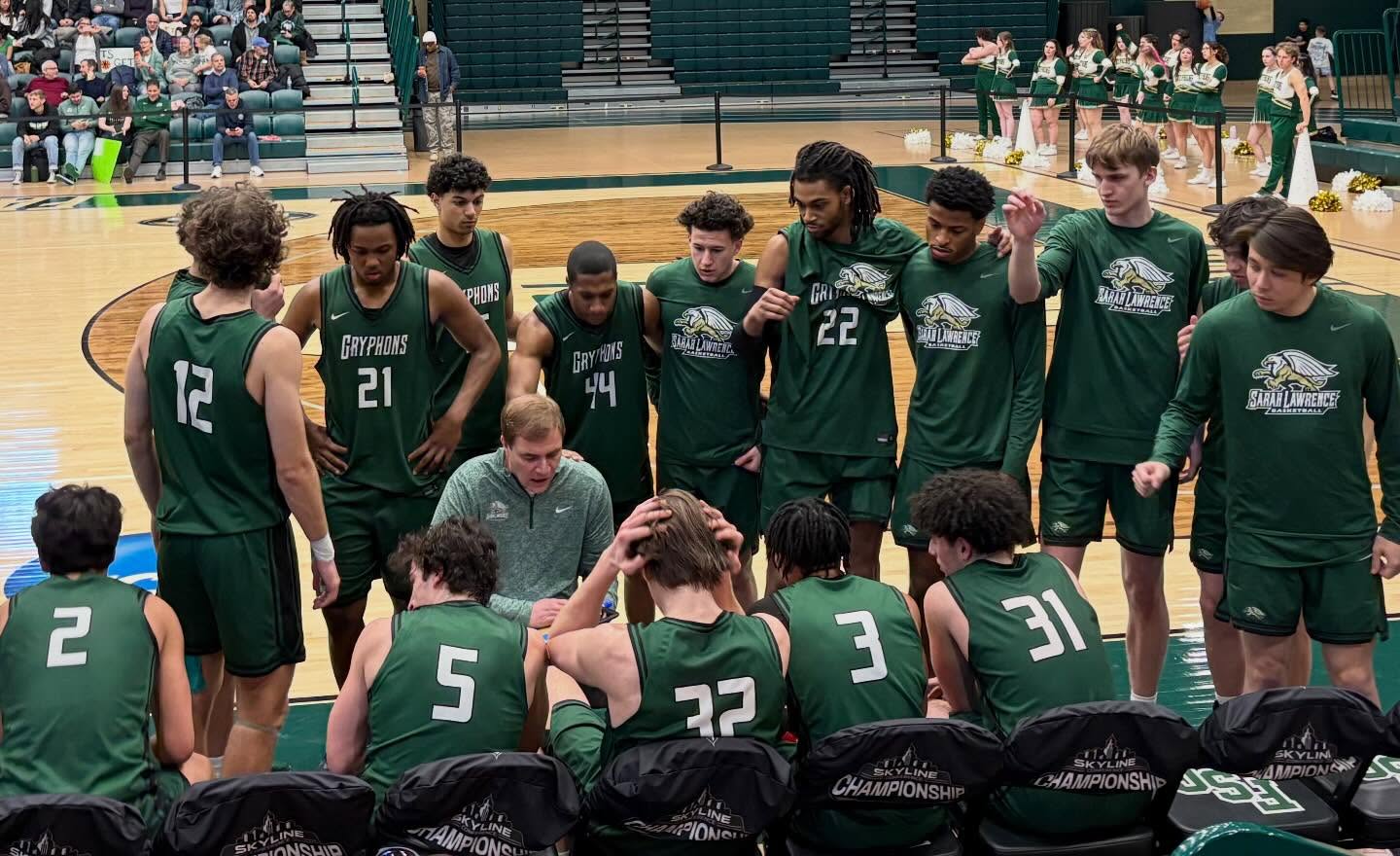 Basketball team in green jerseys huddles around a coach during a timeout on the court, with cheerleaders visible in the background.