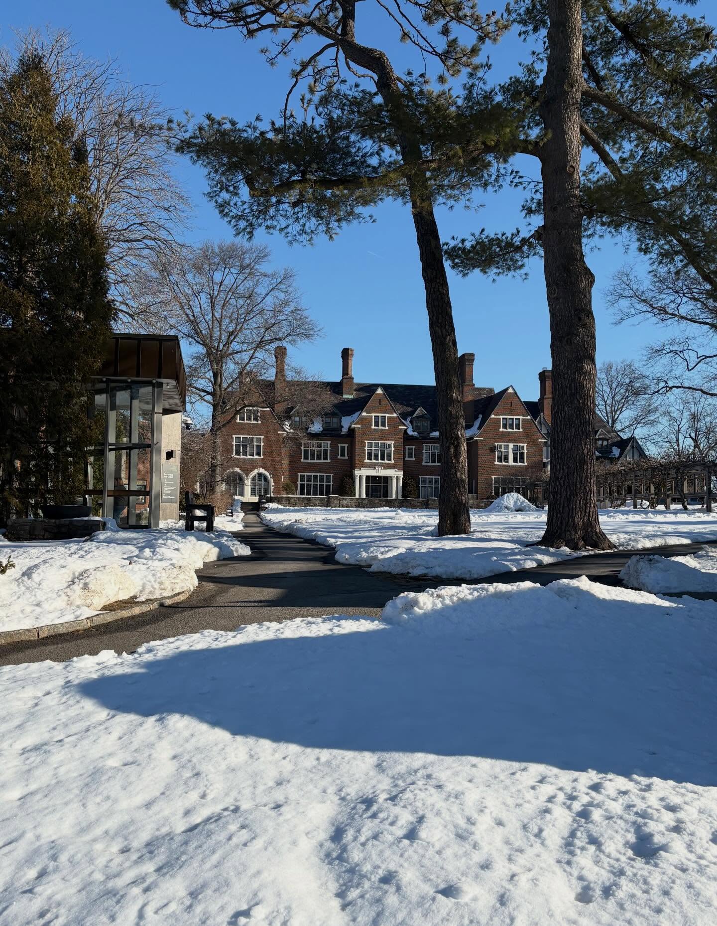 Large brick building with snow-covered grounds and trees under a clear blue sky.