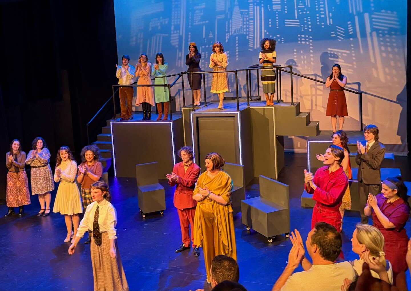 Performers in colorful outfits stand on a multi-level stage, clapping, with a cityscape backdrop and audience members visible in the foreground.
