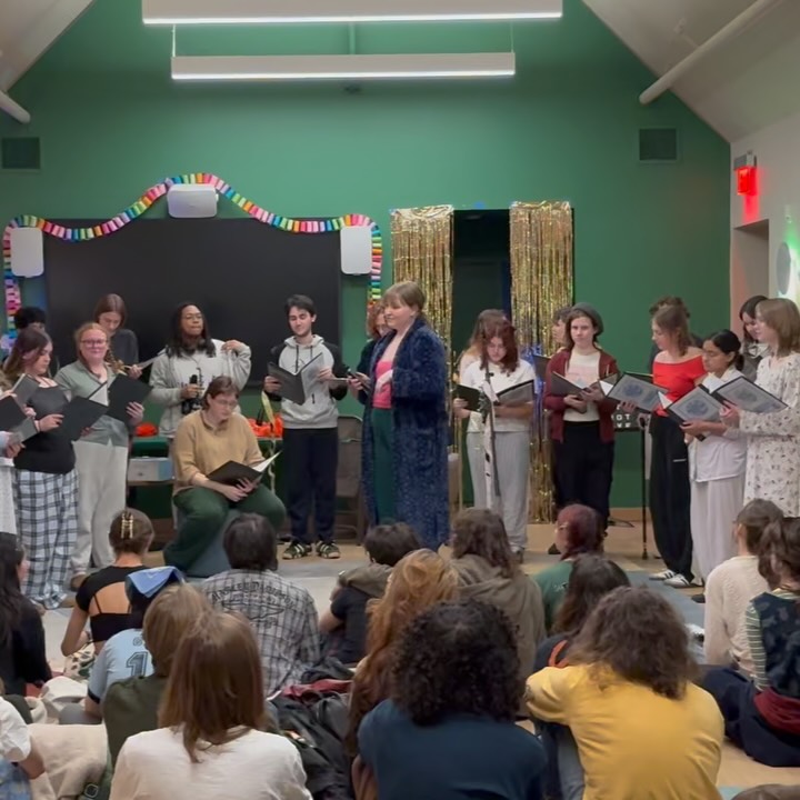 A group of people on stage perform with scripts, facing an audience seated on the floor. The setting includes green walls and colorful decorations.