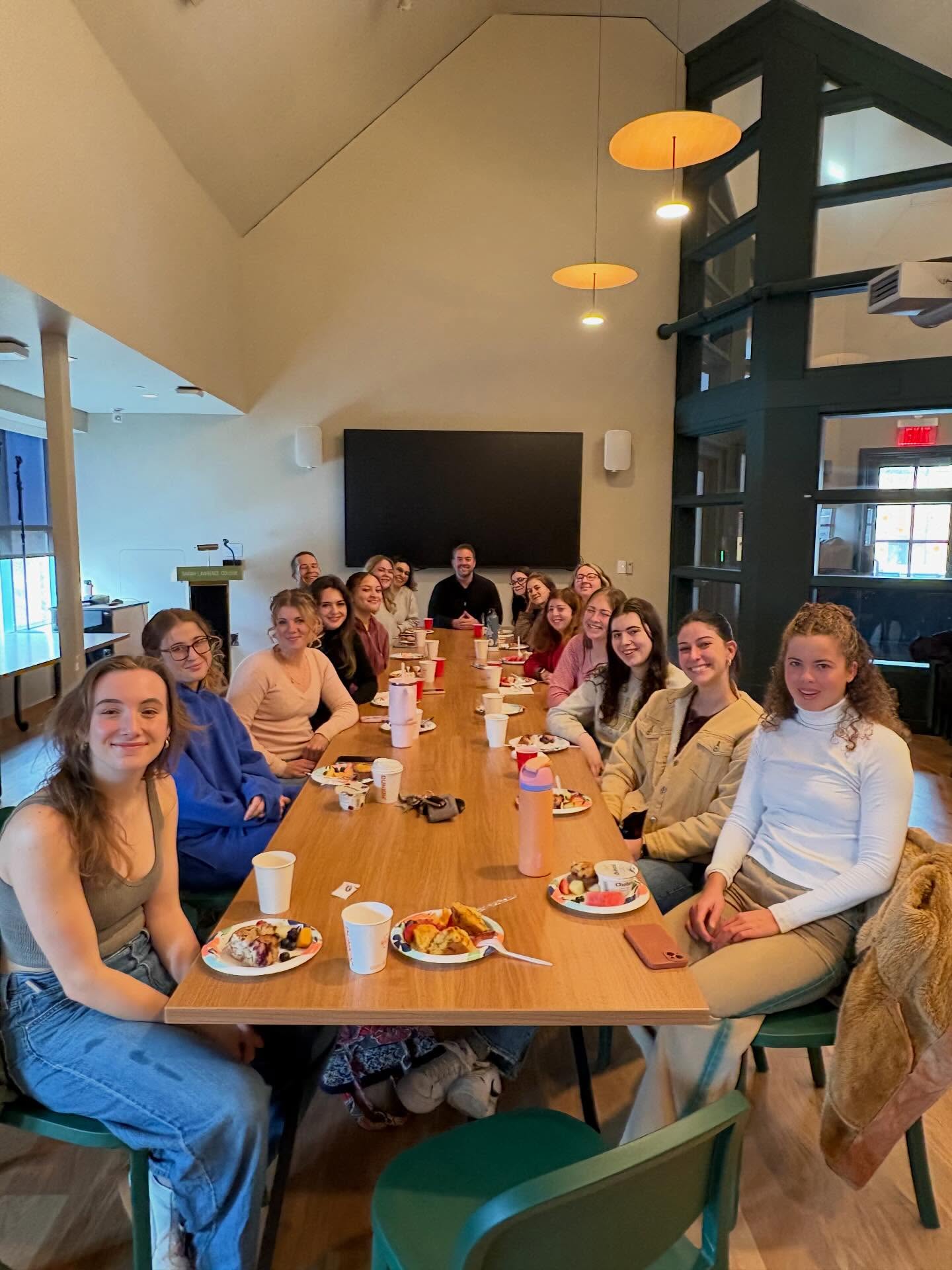 A group of people sit around a long table in a dining area with food and drinks in front of them, smiling at the camera.