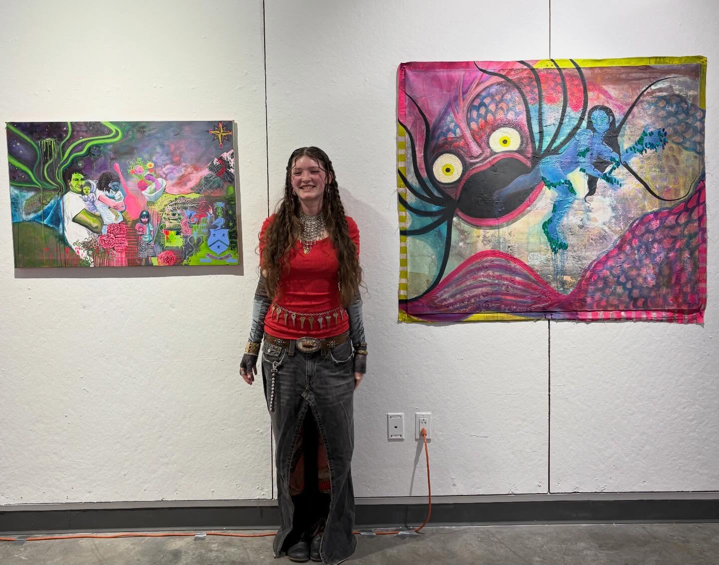Person with long hair smiling, standing between two colorful abstract paintings on a gallery wall.