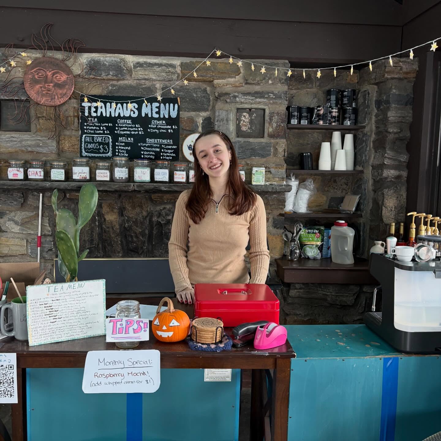 Person standing behind a café counter with a menu board and a display of drinks. A pumpkin and tip jar are on the counter.