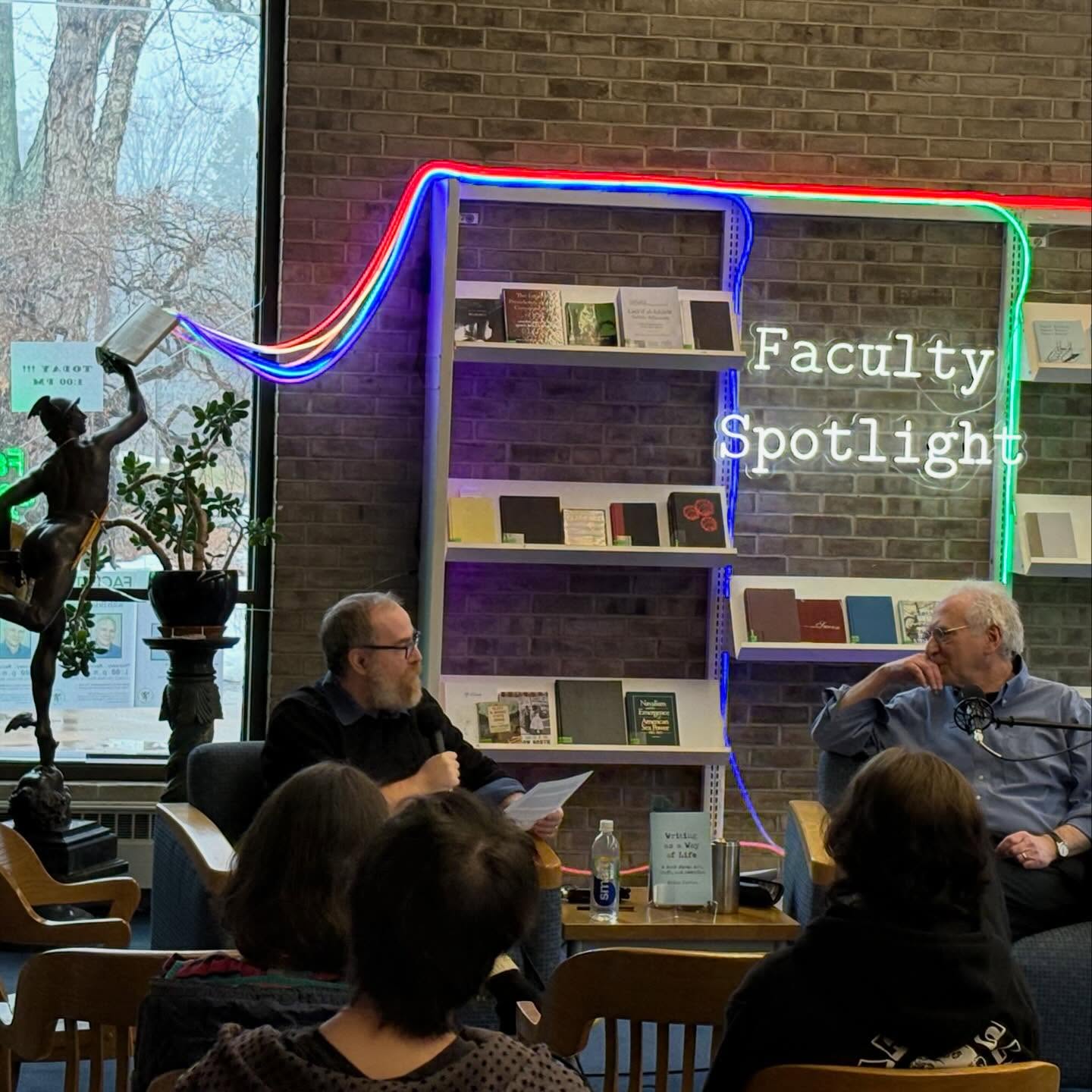 Two people sit and talk in a library setting under a "Faculty Spotlight" neon sign with books displayed in the background.