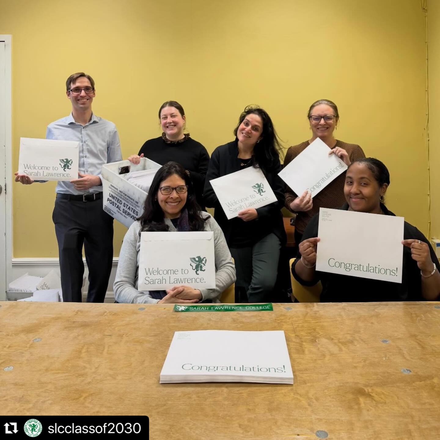 Six people holding Sarah Lawrence College acceptance letters, smiling in a room with yellow walls.