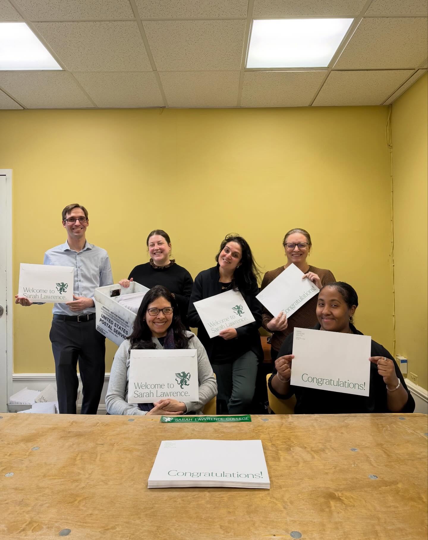 Group of people smiling, holding "Welcome to Sarah Lawrence" and "Congratulations" envelopes in a yellow room.