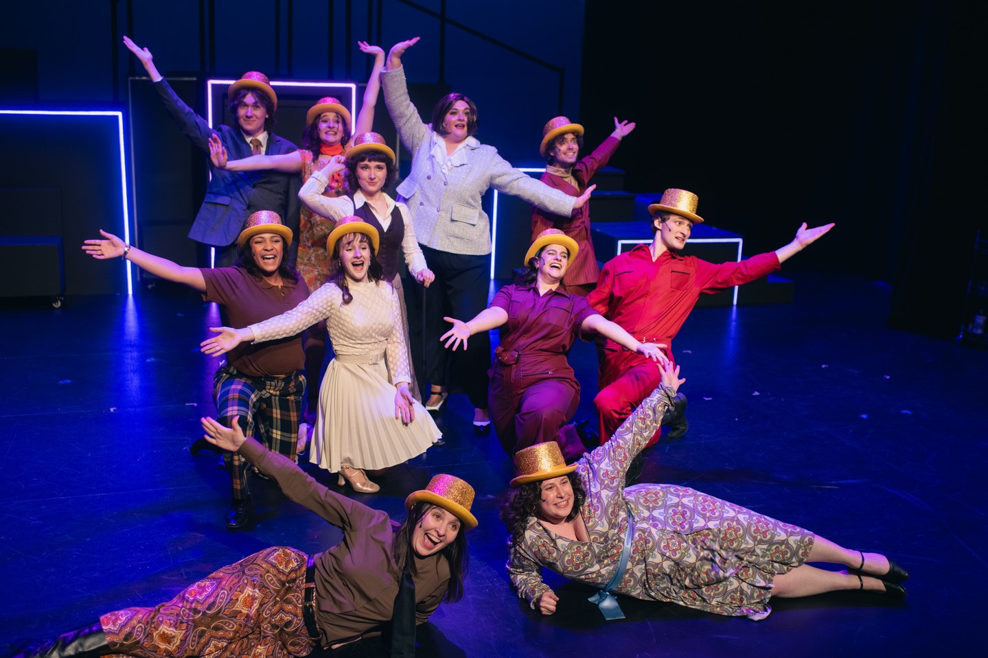 Group of performers on stage wearing yellow hats and colorful outfits, striking playful poses with arms outstretched against a dark backdrop.
