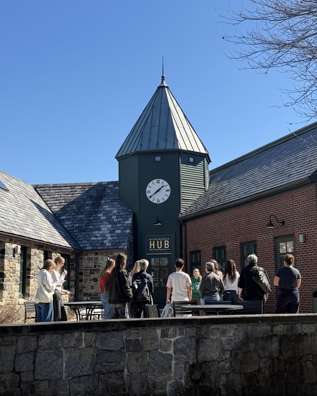 A group of people stand outside a building with a green clock tower and the sign "HUB," under a clear blue sky.