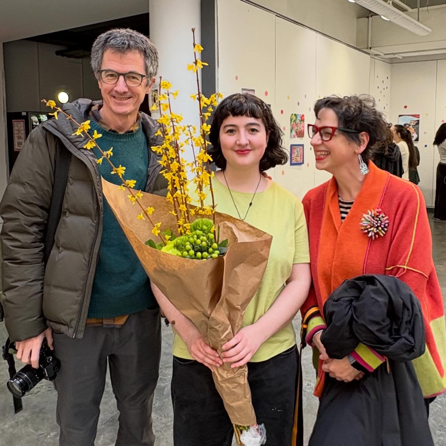 Three people standing together at an art gallery. The center person holds a bouquet of yellow flowers and greenery, smiling at the camera.