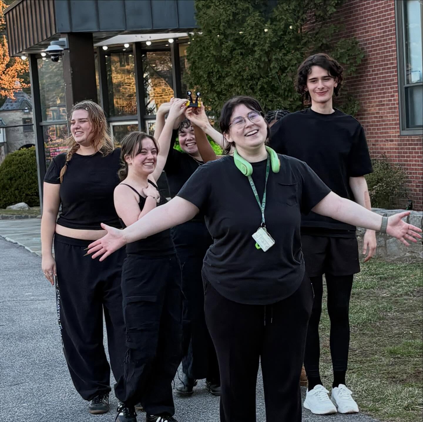 Group of five people in black clothing smiling outdoors, with one person posing playfully in front.