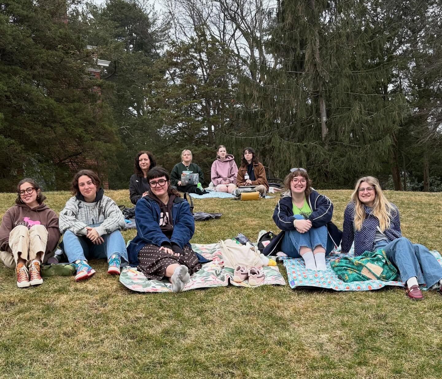 A group of people sitting on blankets on grass, smiling, with trees in the background.