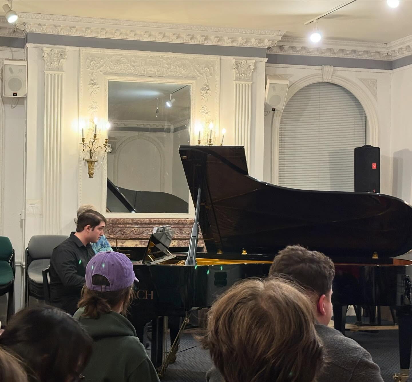 Person playing a grand piano in an ornate room with an audience seated in the foreground.