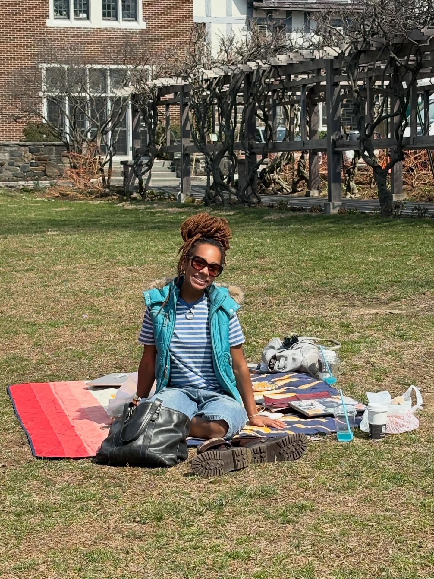 Person sitting on a blanket in a park with sunglasses, wearing a striped shirt and vest. Background has buildings and a pergola.