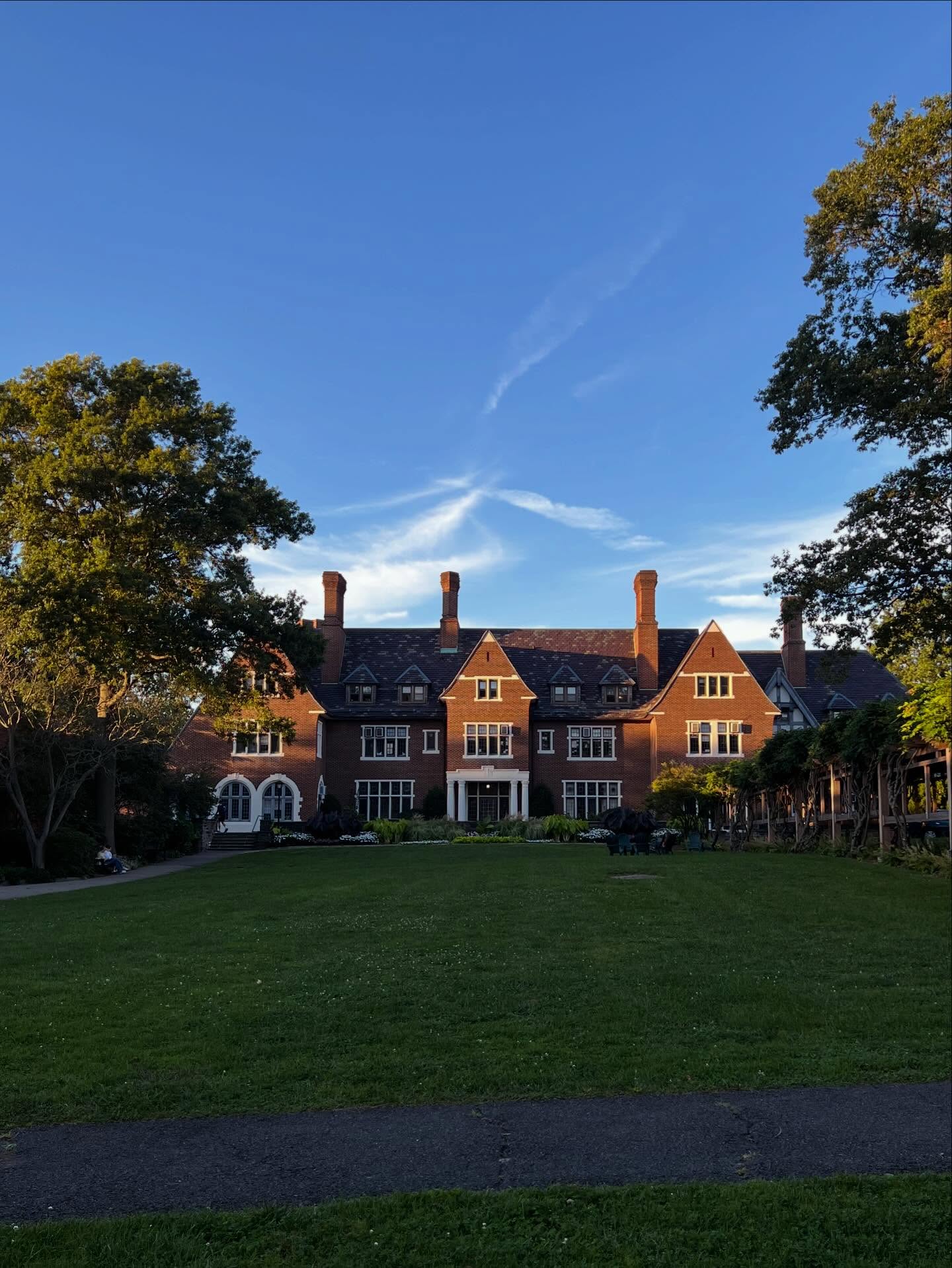 A large brick mansion with multiple chimneys, surrounded by trees, under a clear blue sky. A well-kept lawn and pathway lead to the entrance.