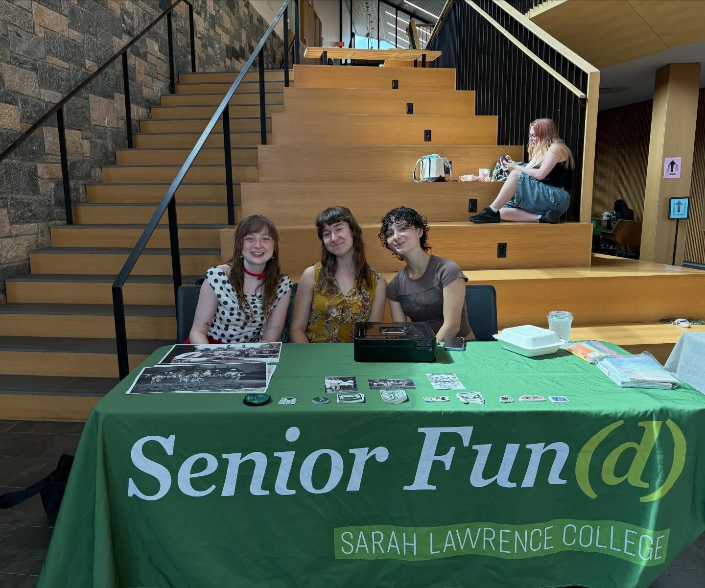 Three people sit at a "Senior Fun(d)" booth with stickers and items. Another person is seated on steps nearby in the background.