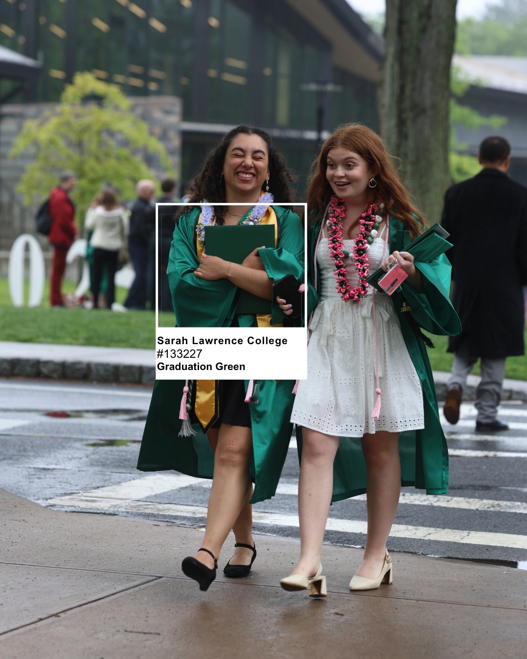 Two graduates in green gowns, holding diplomas, walking on a rainy day. They are smiling, with school buildings and people in the background.