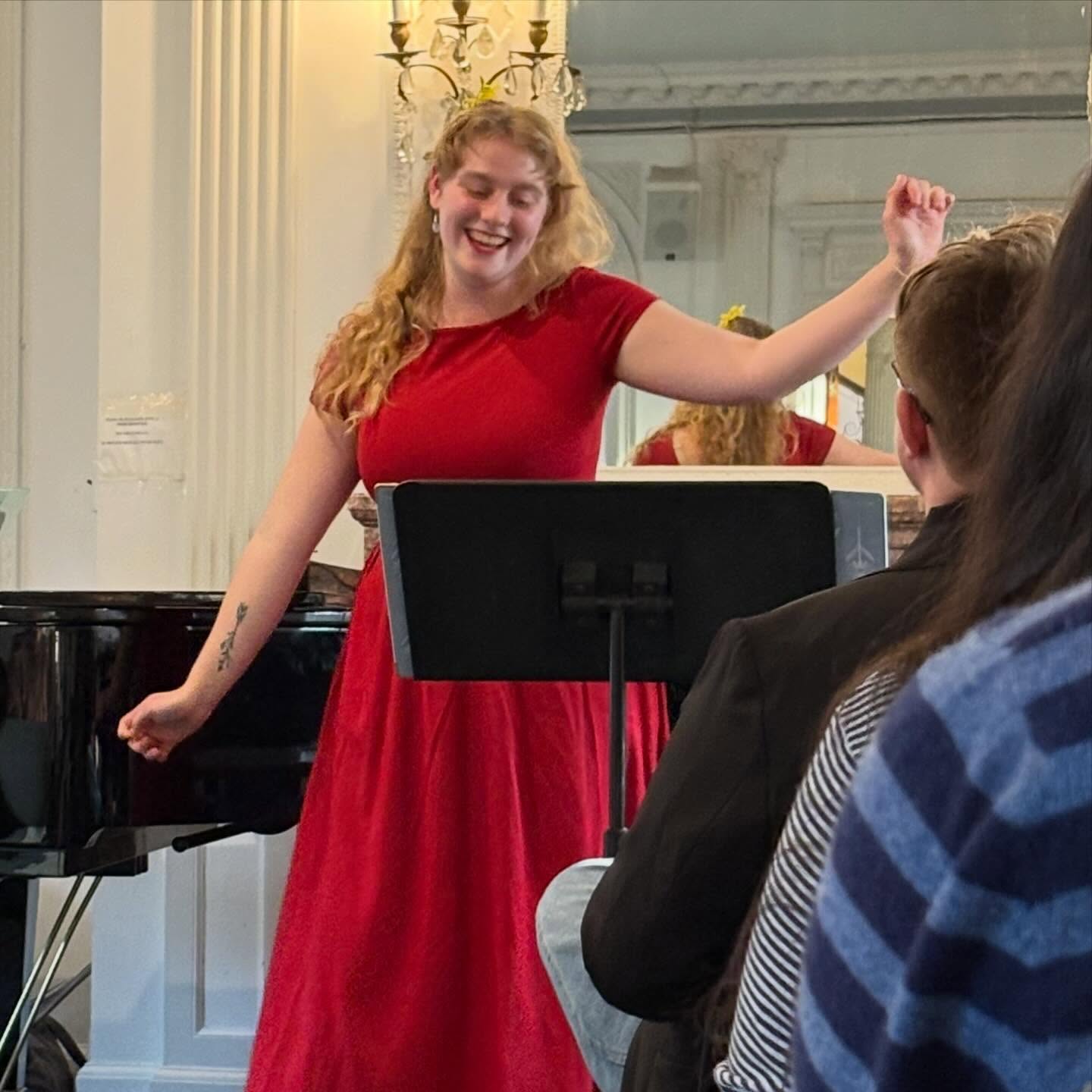Person in red dress performing in front of a music stand, with a piano and audience in a room with elegant decor.