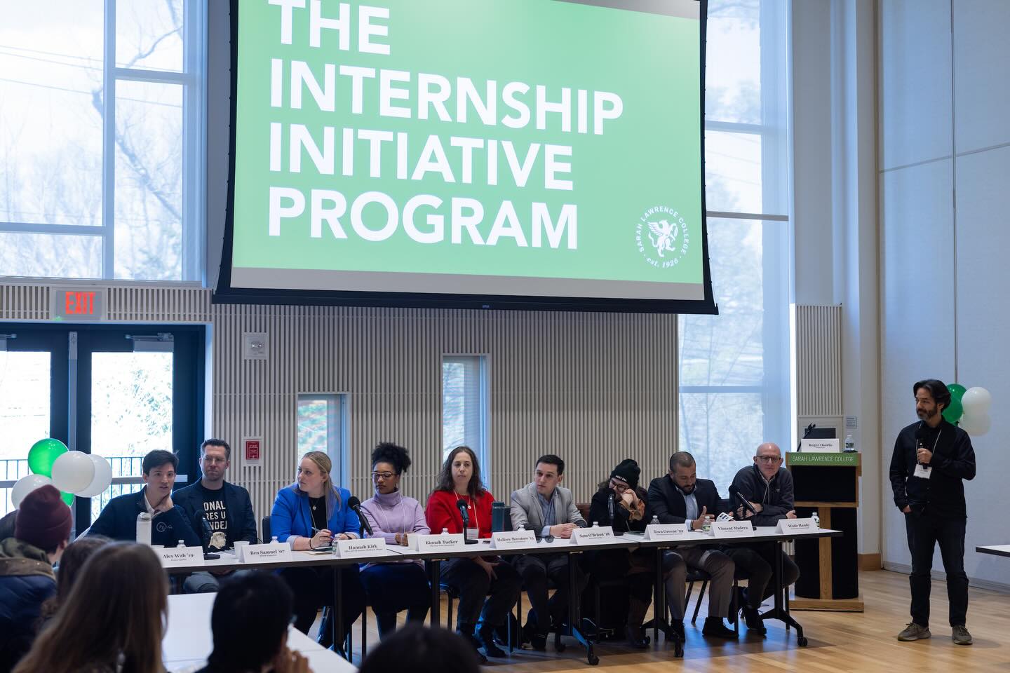 Panel discussion with multiple individuals seated at a table and one standing, under a sign reading "The Internship Initiative Program."